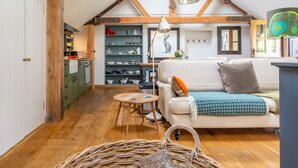 A cozy living room with a sofa, a small round table, a wicker basket, and a bookshelf with various items. - Old Church Hall, Glandford, Norfolk (Glandford)
