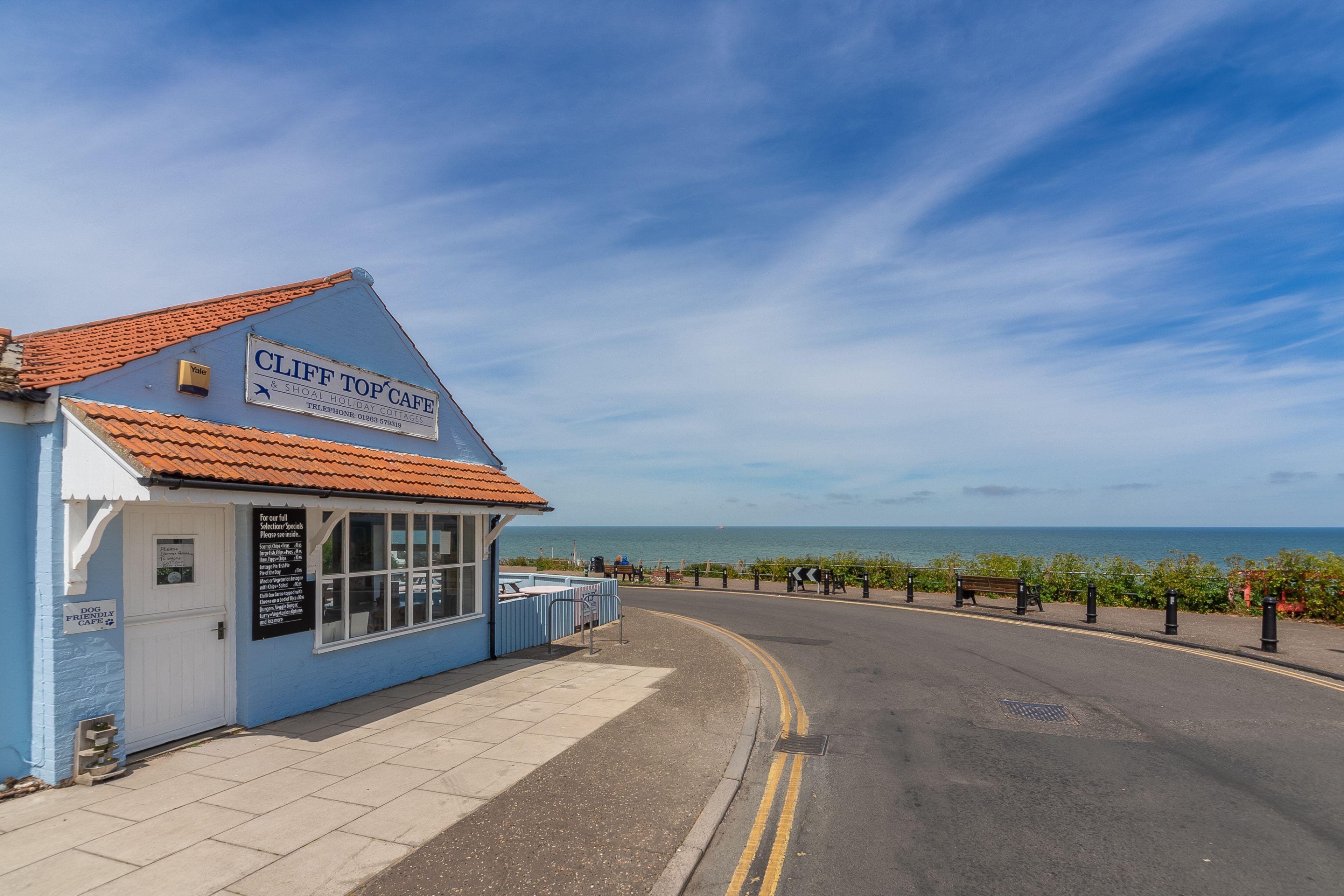 A coastal cafe with a blue exterior and a tiled roof, located beside a road with a view of the sea.