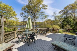 A deck with outdoor furniture, a table, and chairs, surrounded by trees and a view of a lake. - Swan Dive - Waterfront Retreat in Cataumet RPLO032 (Bourne)