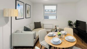 Ein modernes Wohnzimmer mit einer Couch, einem Esstisch für vier Personen, einem Fernseher und gerahmten Kunstwerken an der Wand.