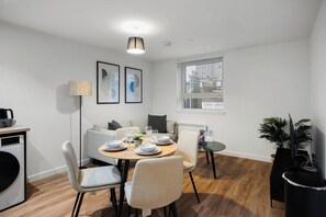 Een moderne eetruimte met een ronde tafel gedekt voor vier personen, een bank, een klein bijzettafeltje en een tv-meubel met een plant.