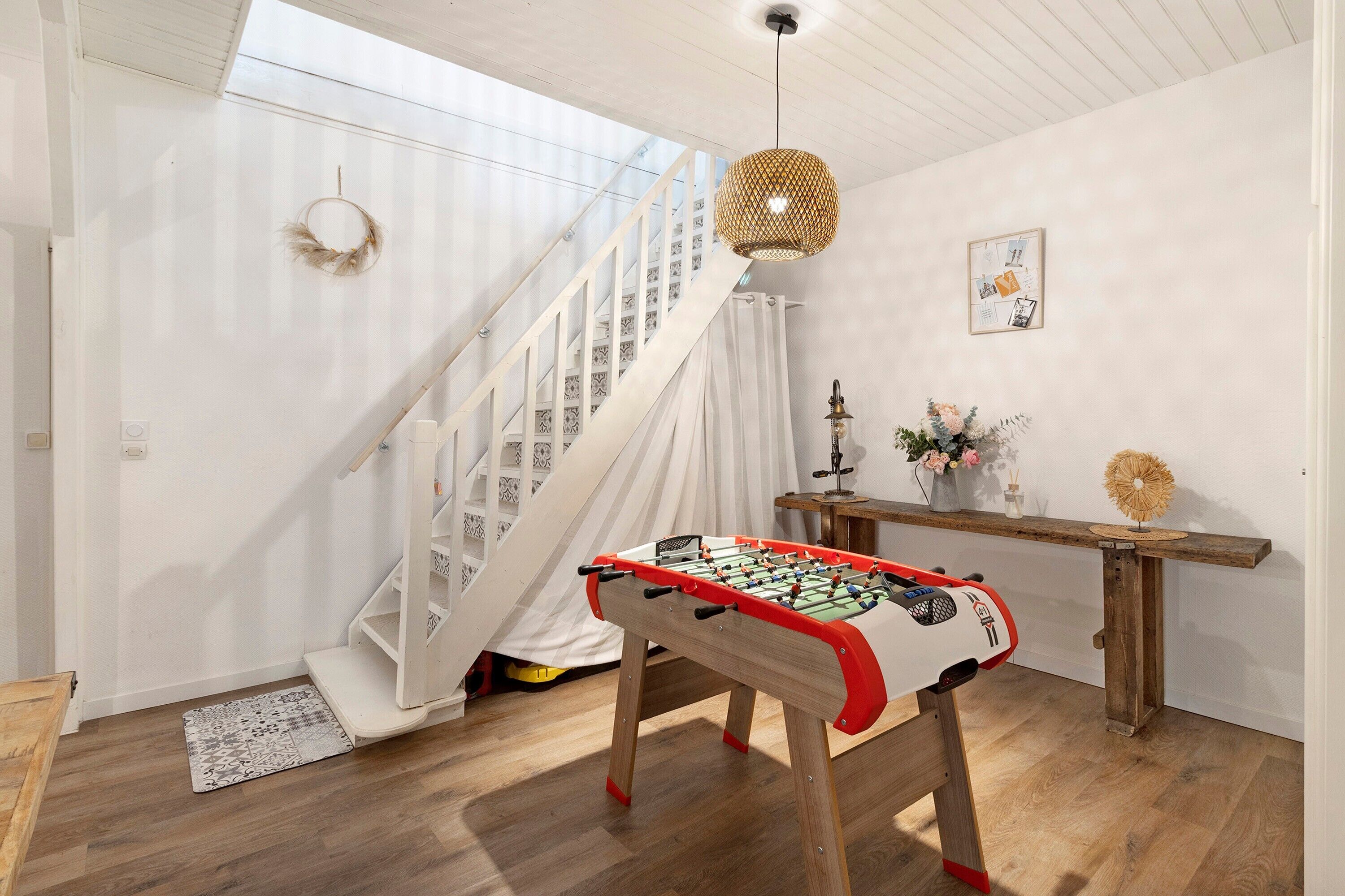Une pièce avec une table de baby-foot, un escalier, un luminaire suspendu et un tableau accroché au mur.
