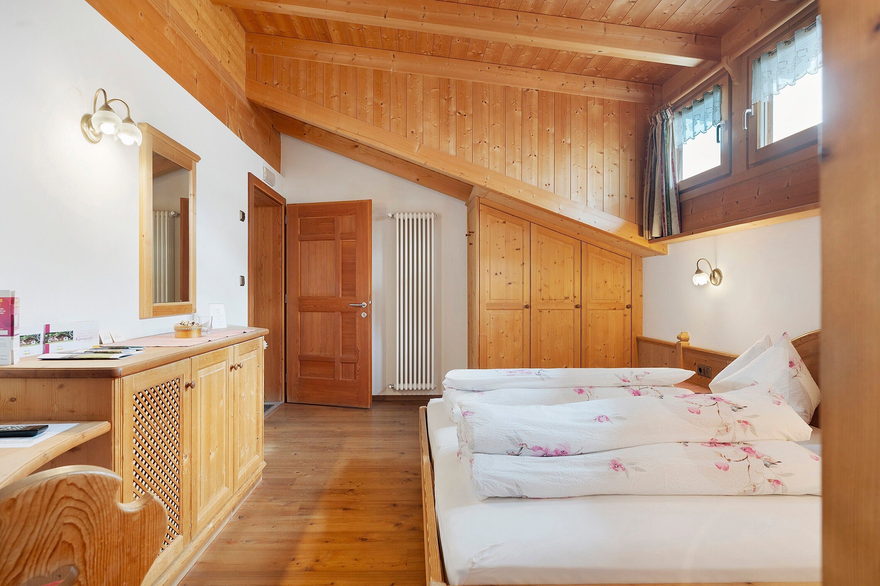 Une chambre avec un plafond en bois, un lit recouvert d’une literie à motifs floraux, un bureau en bois et un radiateur.