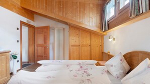 A bedroom with wooden ceiling beams, a bed with floral bedding, a wooden wardrobe, and a bedside table with a lamp.