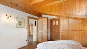 A bedroom with wooden ceiling beams, a bed with floral bedding, wooden wardrobes, and a hallway leading to another room. - Apartment Margerite 21 with Mountain View and Wi-Fi (Santa Valburga)