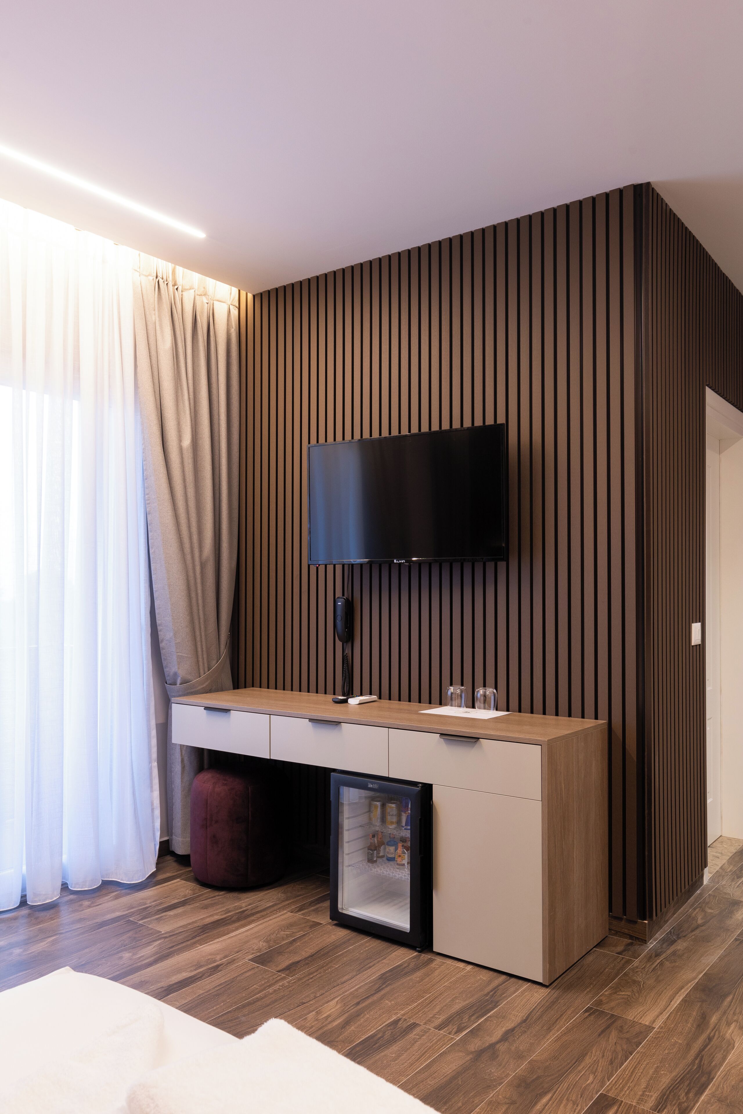 A modern room with a wooden desk, a flat-screen TV mounted on the wall, a mini-fridge, and a red ottoman.