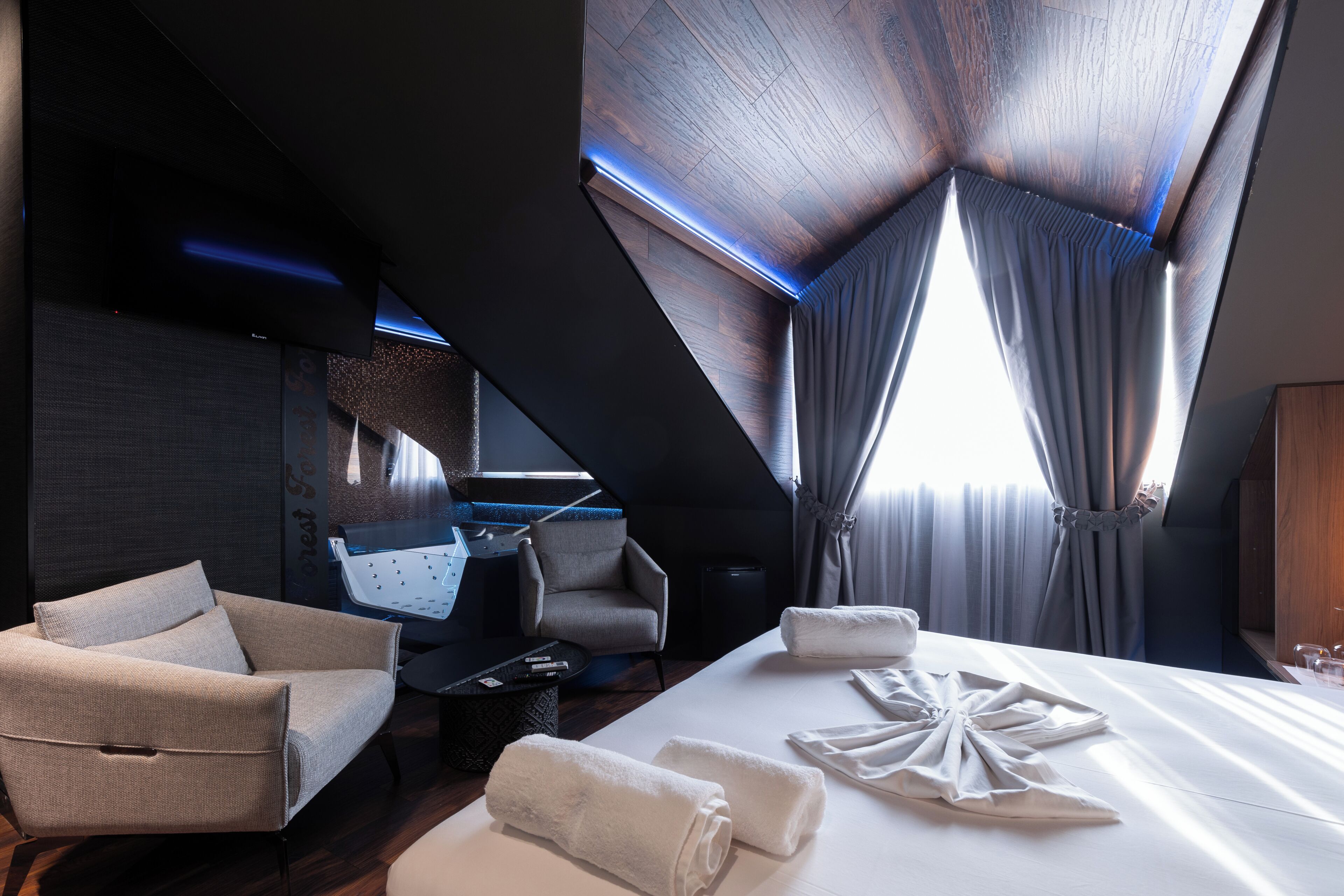 A modern hotel room with a large bed, a seating area with a sofa and armchairs, a small table, and a view of the outdoors.