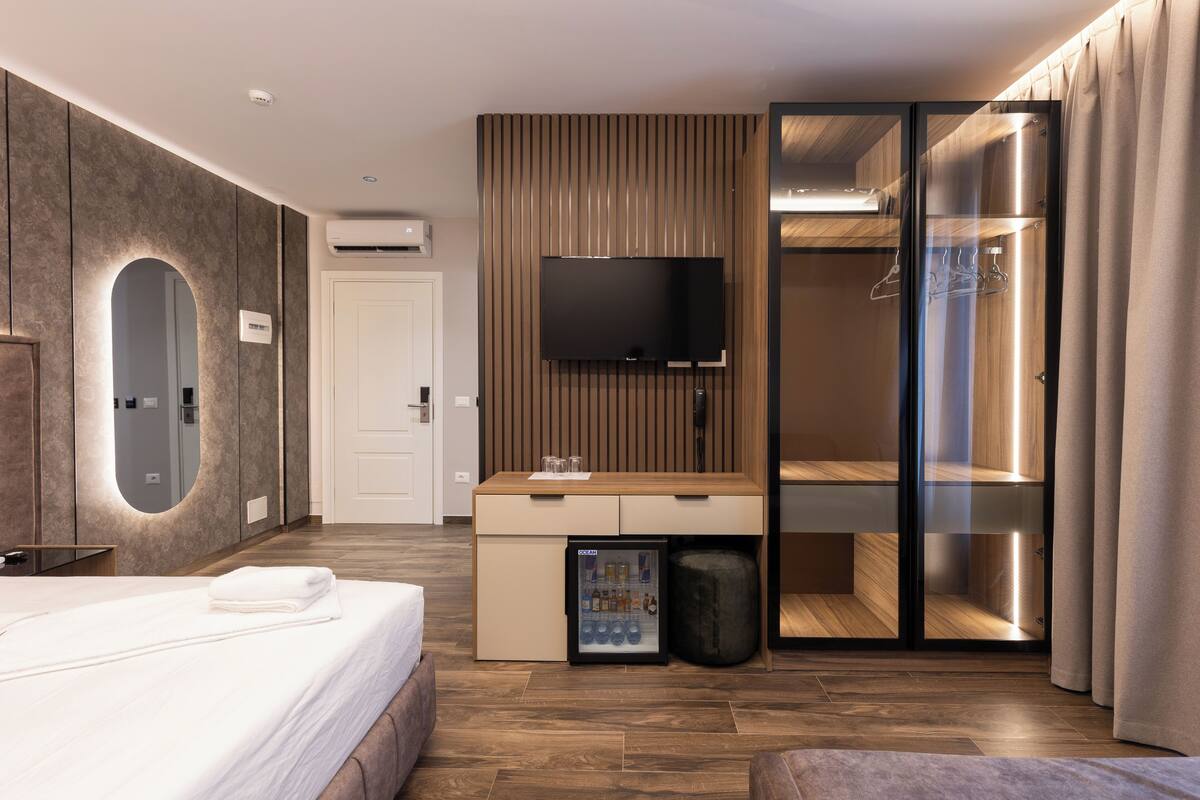 Moderni hotellihuone, jossa on sänky, televisio, minibaari, peili ja liukuovilla varustettu vaatekaappi.