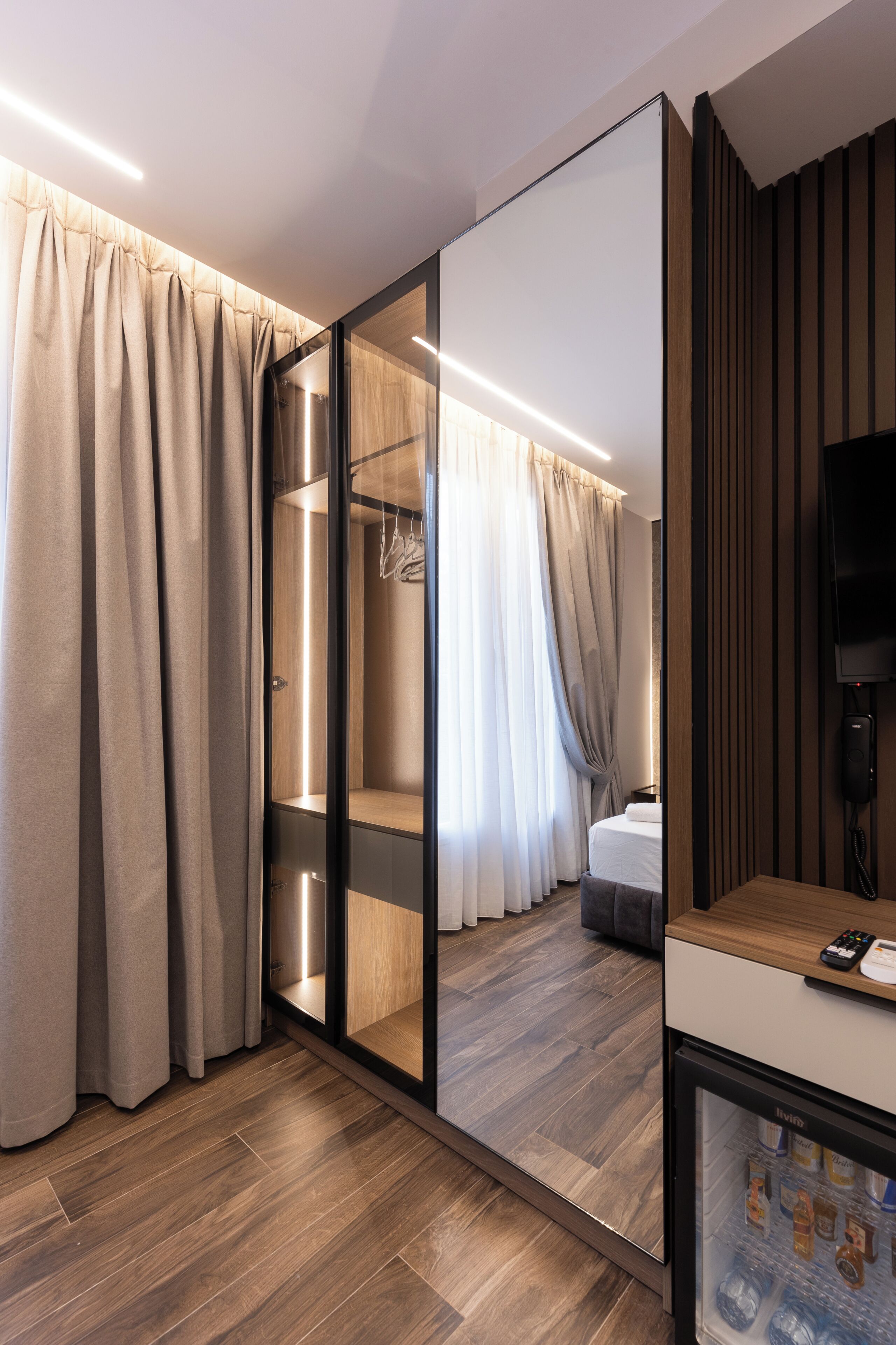 Una camera d'albergo moderna con un grande specchio, pavimento in legno, un armadio e un frigobar.