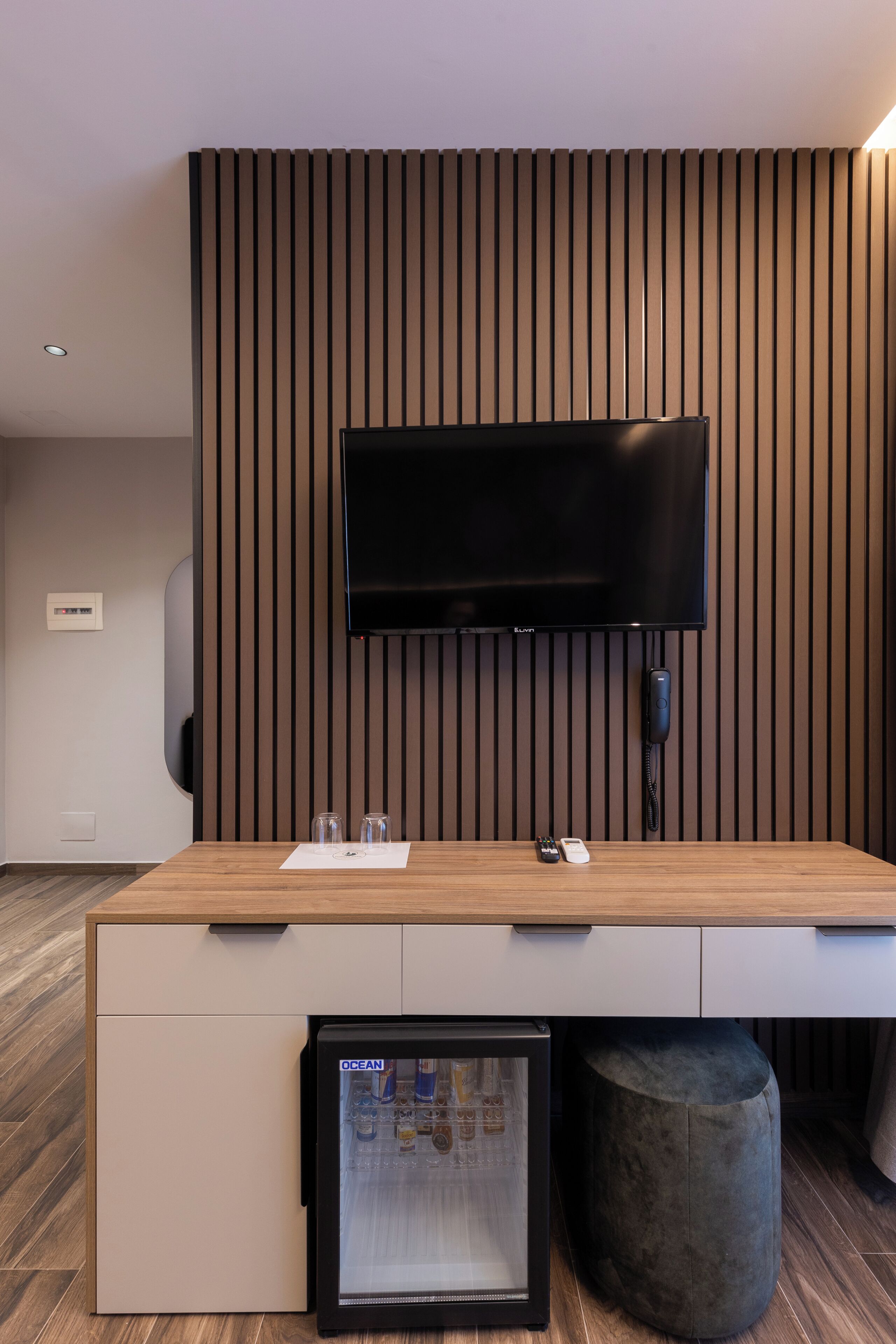 A modern hotel room with a flat-screen TV mounted on a wooden wall, a minibar, and a small refrigerator.