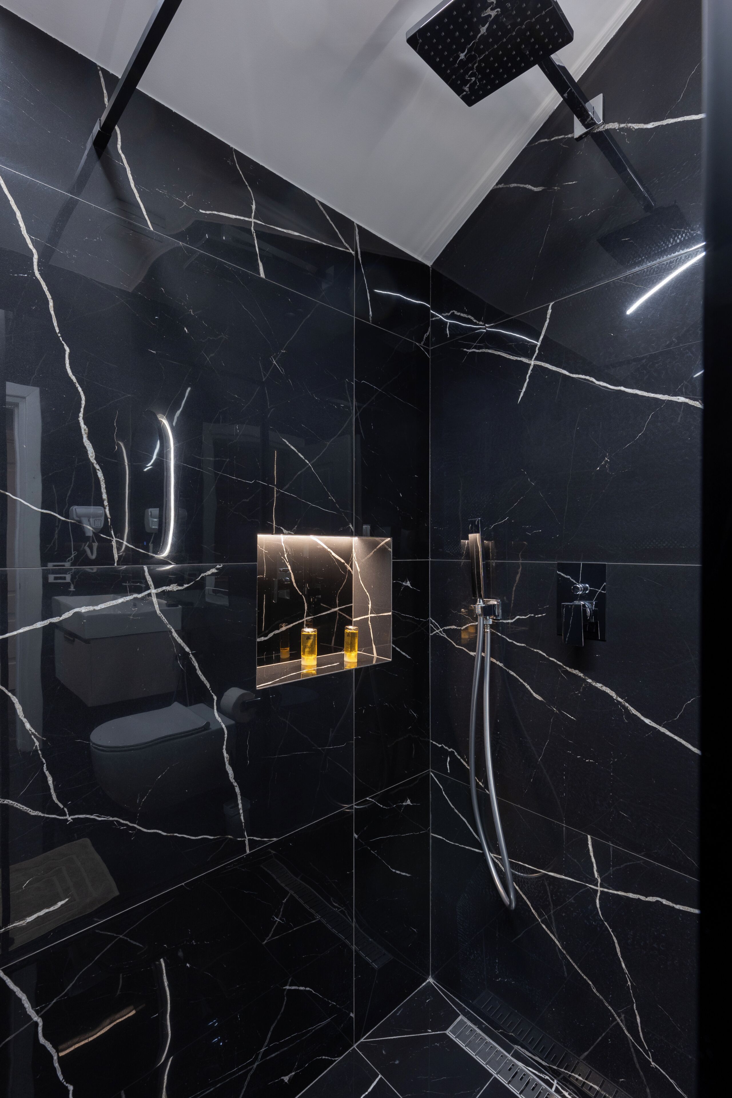 Un baño moderno con paredes de mármol negro, cabina de ducha de vidrio y espejo integrado.