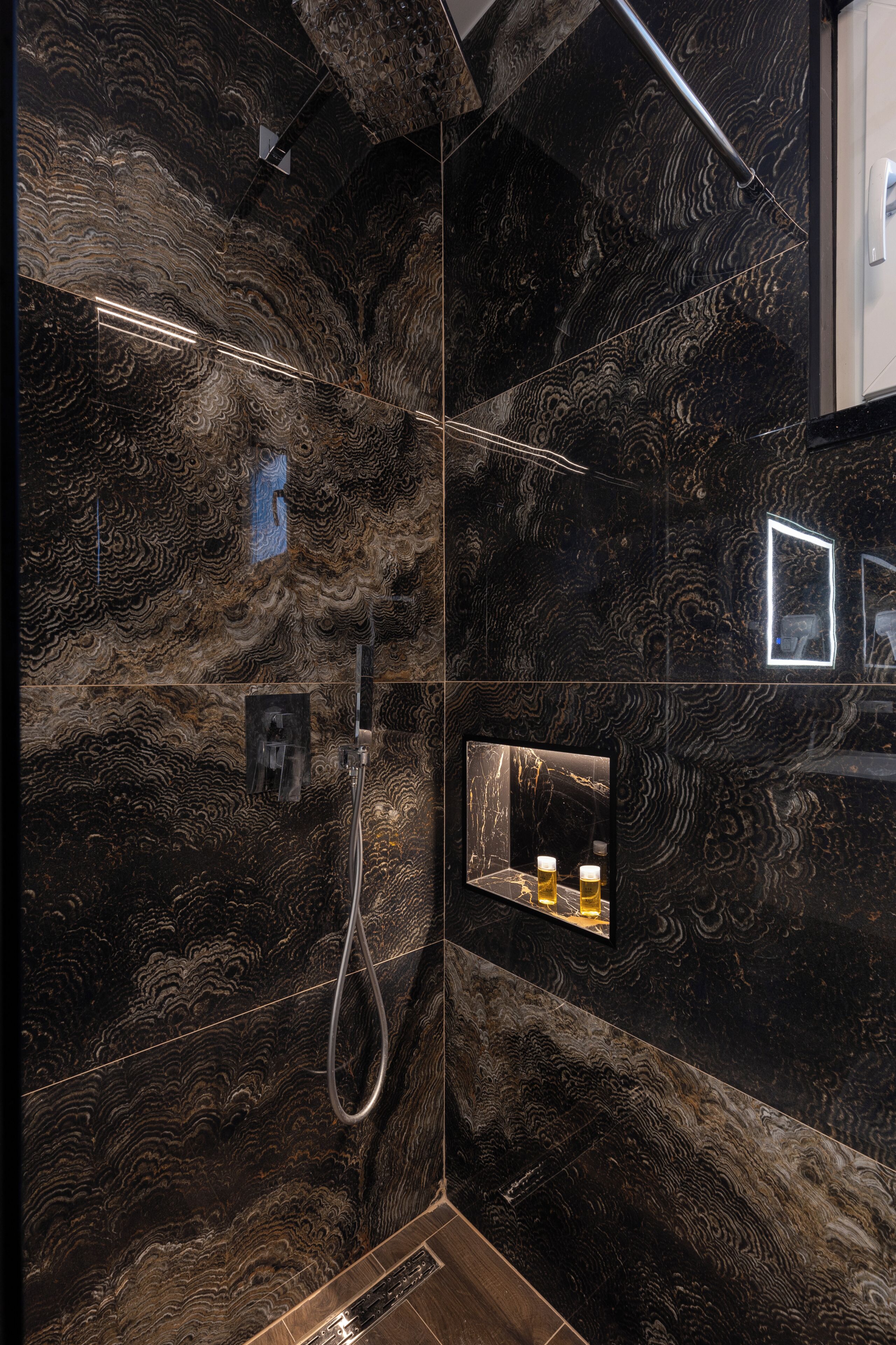 Un baño moderno con pared de mármol negro, zona de ducha y un pequeño espejo con luces.