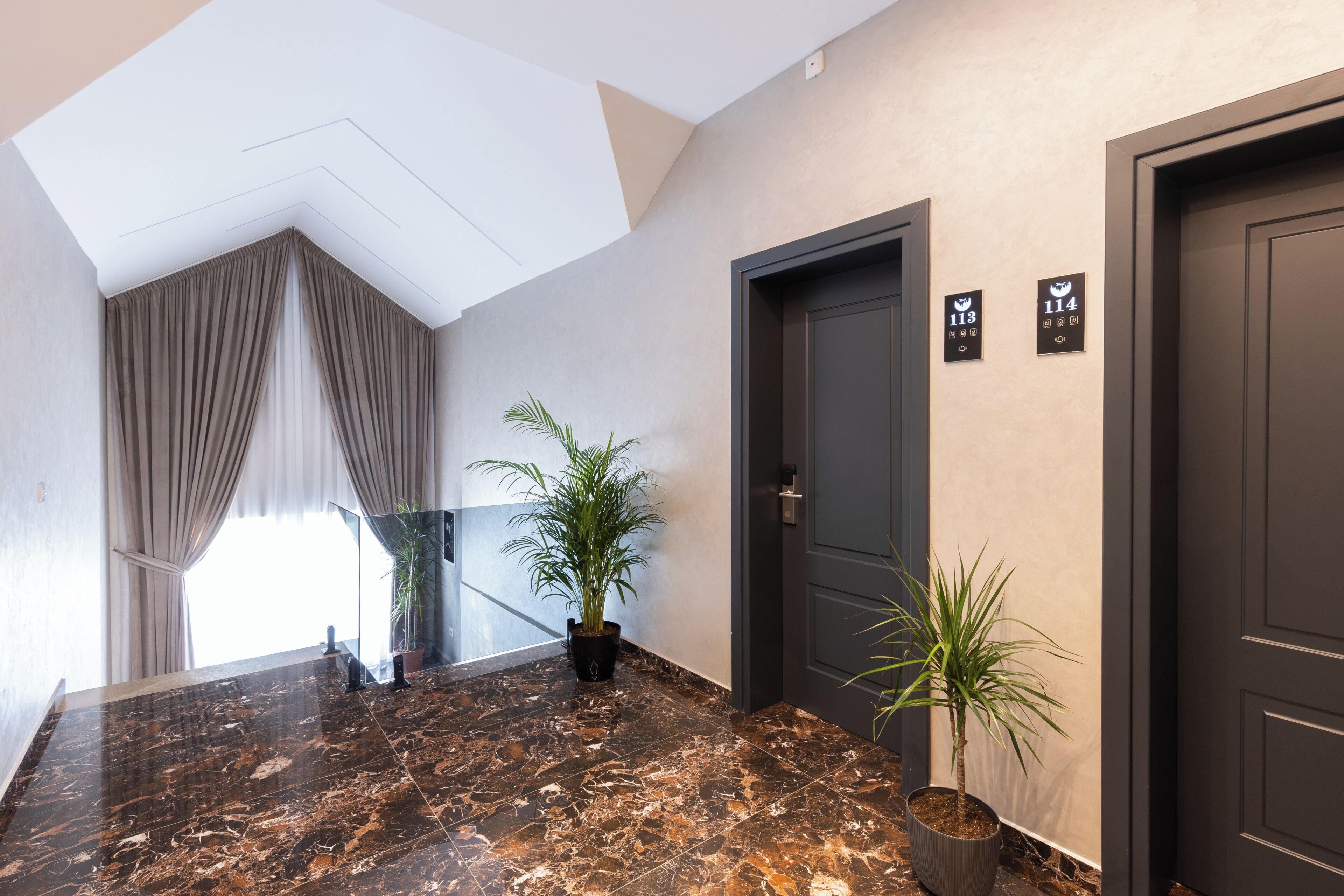A hotel hallway with a marble floor, two potted plants, a large window with curtains, and doors marked with room numbers.