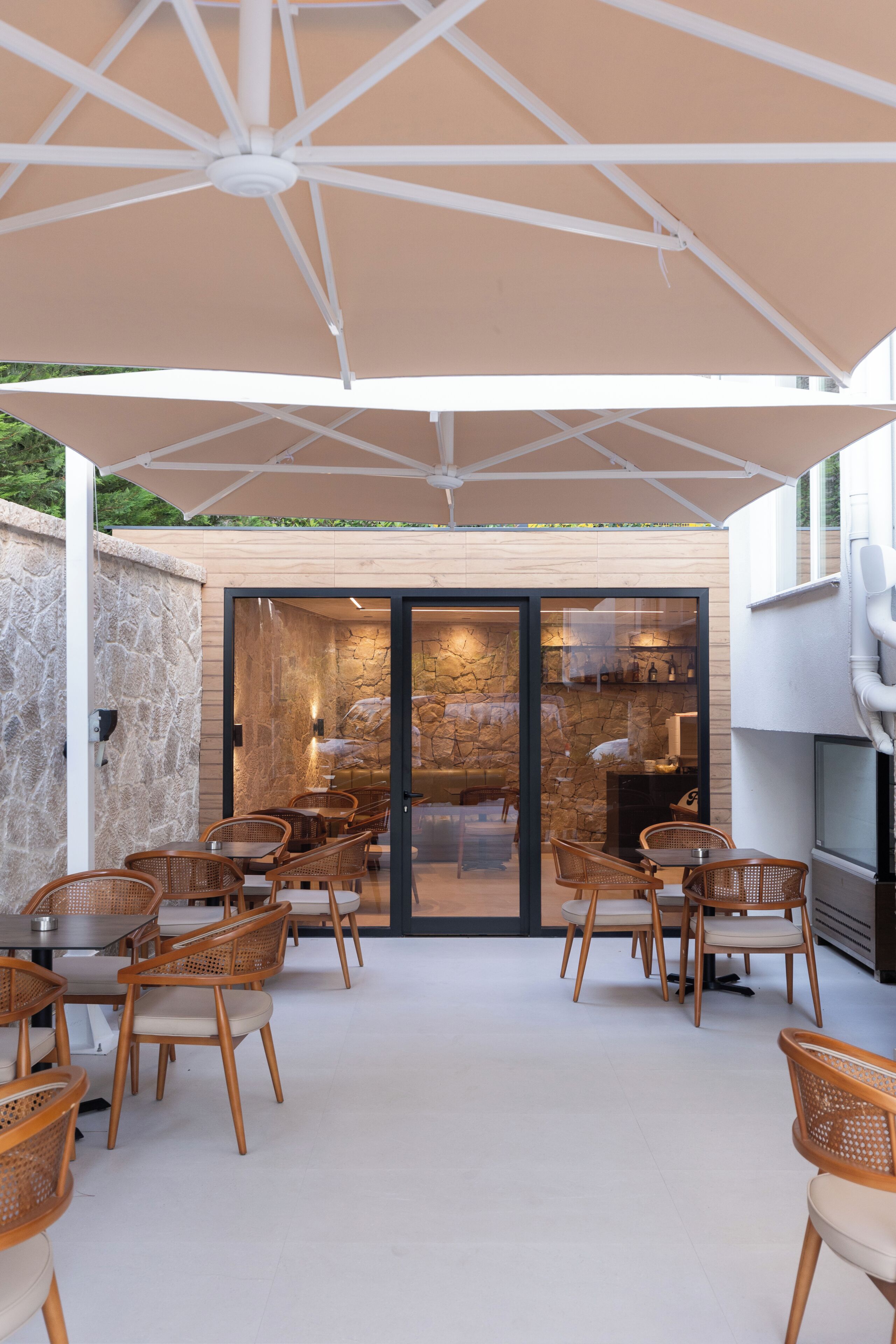 An outdoor patio with wicker chairs and a large umbrella, a stone wall, and a glass door leading to an interior space with a fireplace.