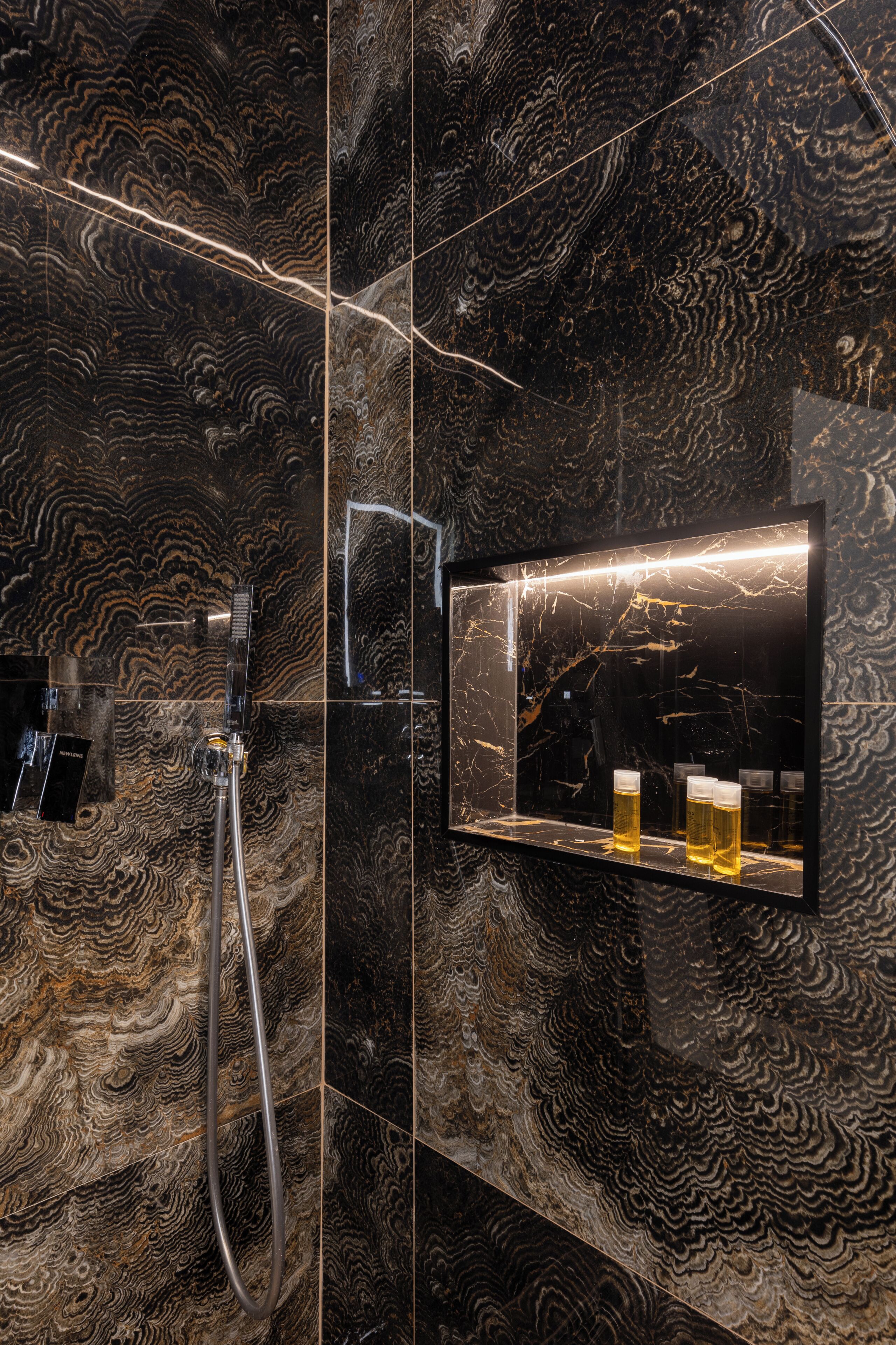 A bathroom with a marble wall, a glass shower enclosure, and a mirror with a shelf holding glasses.