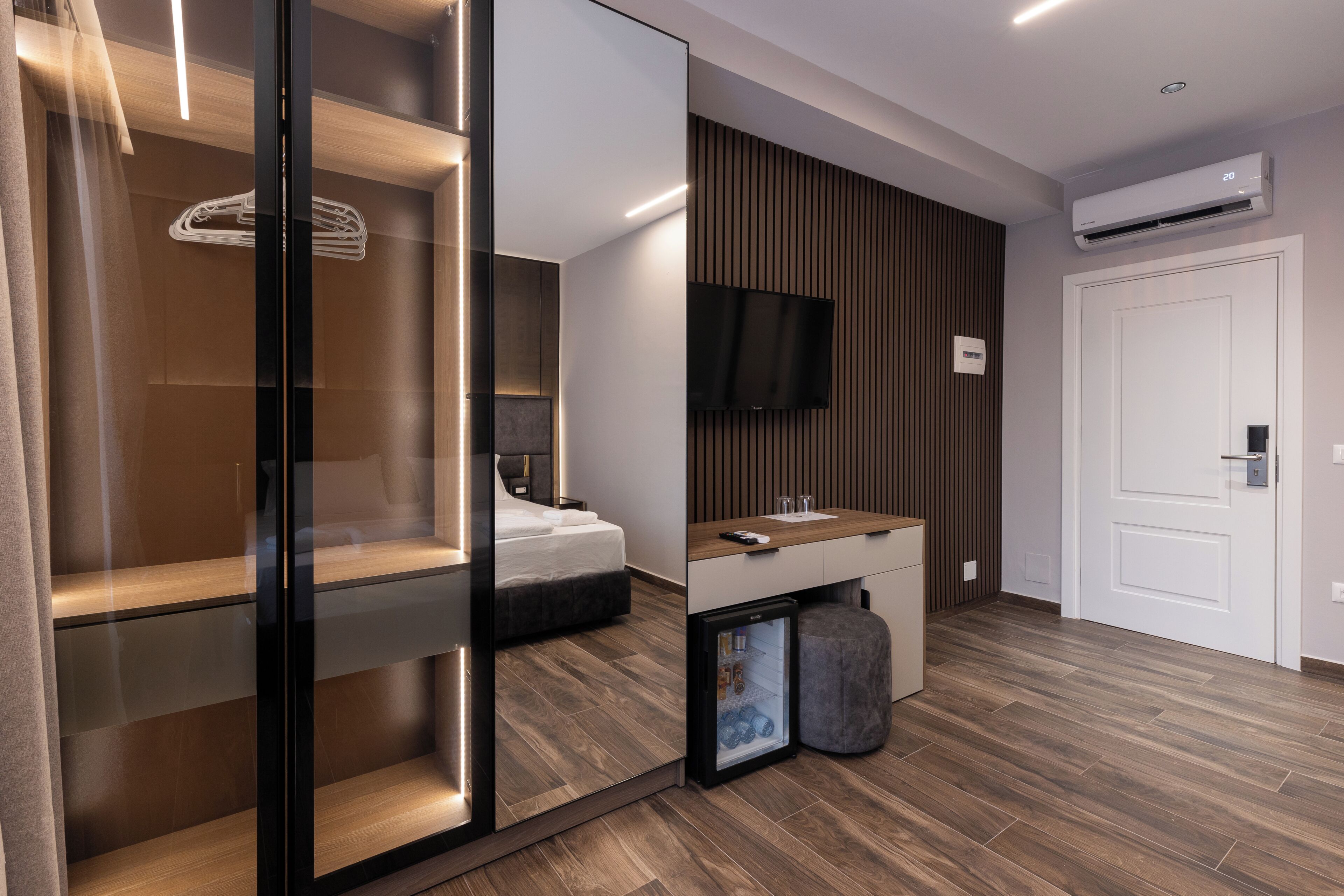 A modern hotel room with a bed, a built-in wardrobe, a small kitchenette, a television, and an air conditioning unit.