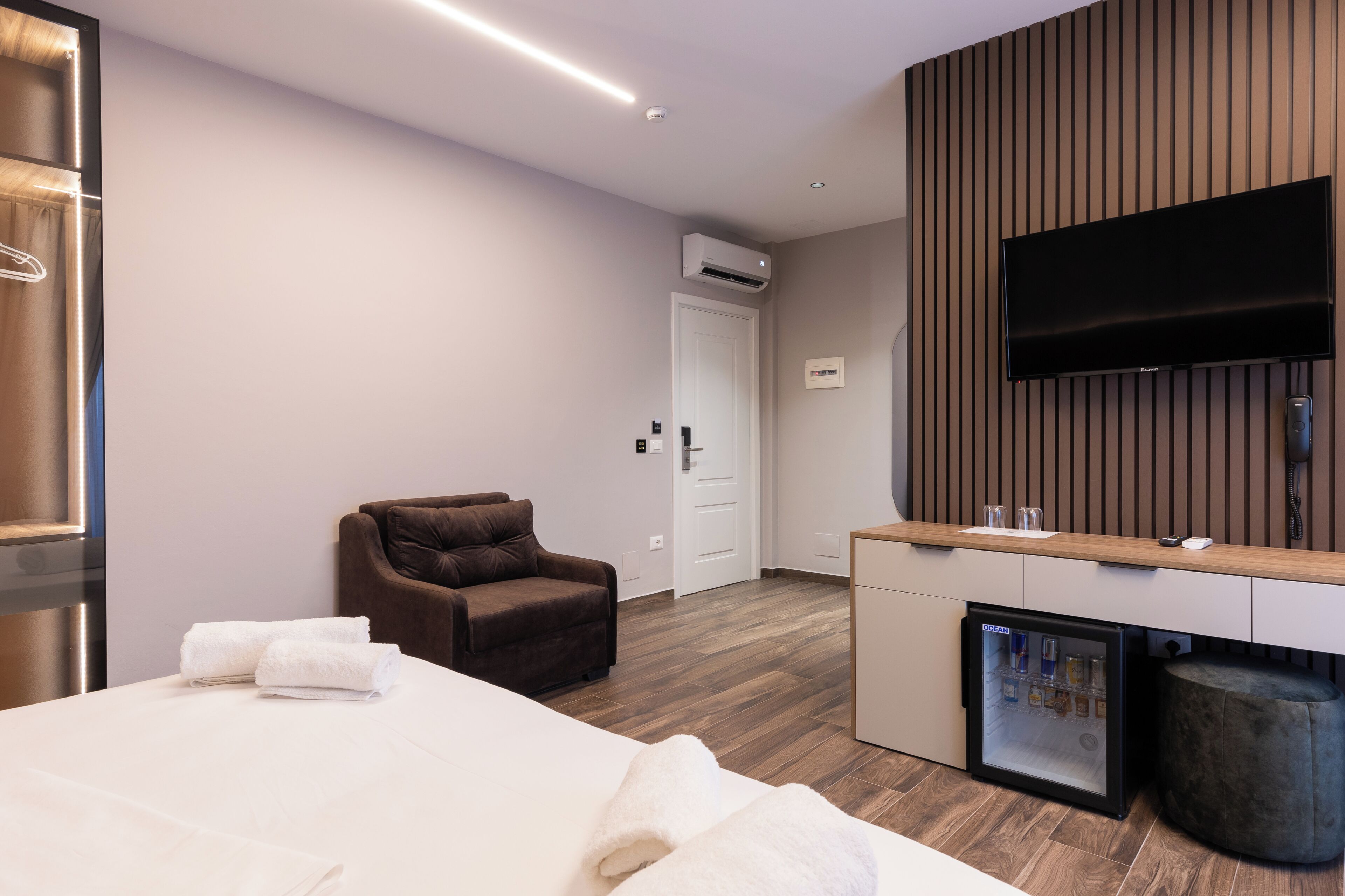 Ein modernes Hotelzimmer mit Bett, Sitzecke, Minibar und einem an einer Holzpaneelwand montierten Flachbildfernseher.