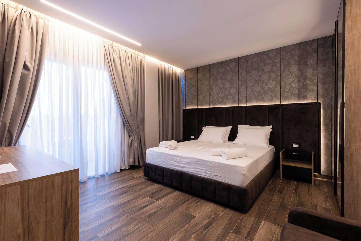 Moderni hotellihuone, jossa on suuri sänky, työpöytä ja näkymä ulos ohuiden verhojen läpi.
