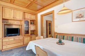 Ein Zimmer mit Holzmöbeln, einem Fernseher, einem Esstisch mit weißem Tischlauf und einer Bank mit farbigem Kissen.