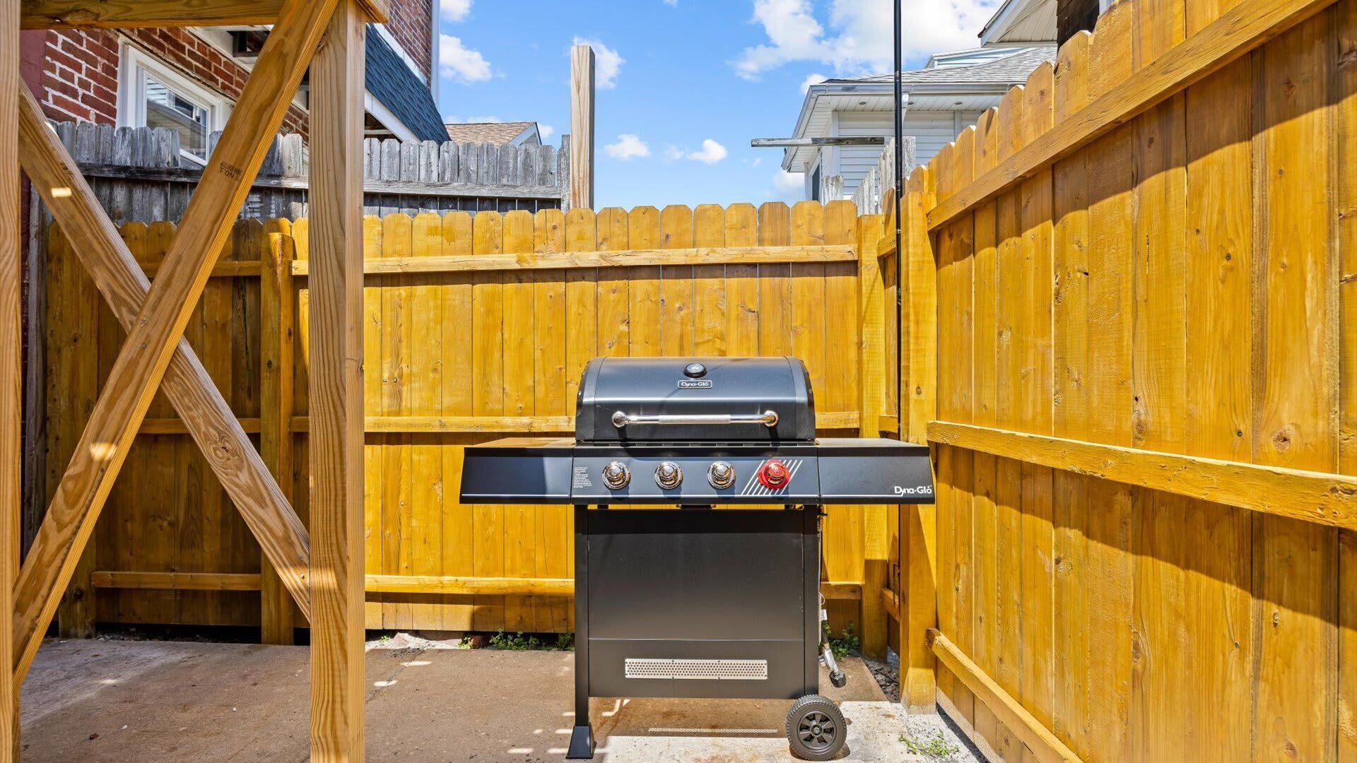 Patio trasero con una parrilla, valla de madera y una casa al fondo.