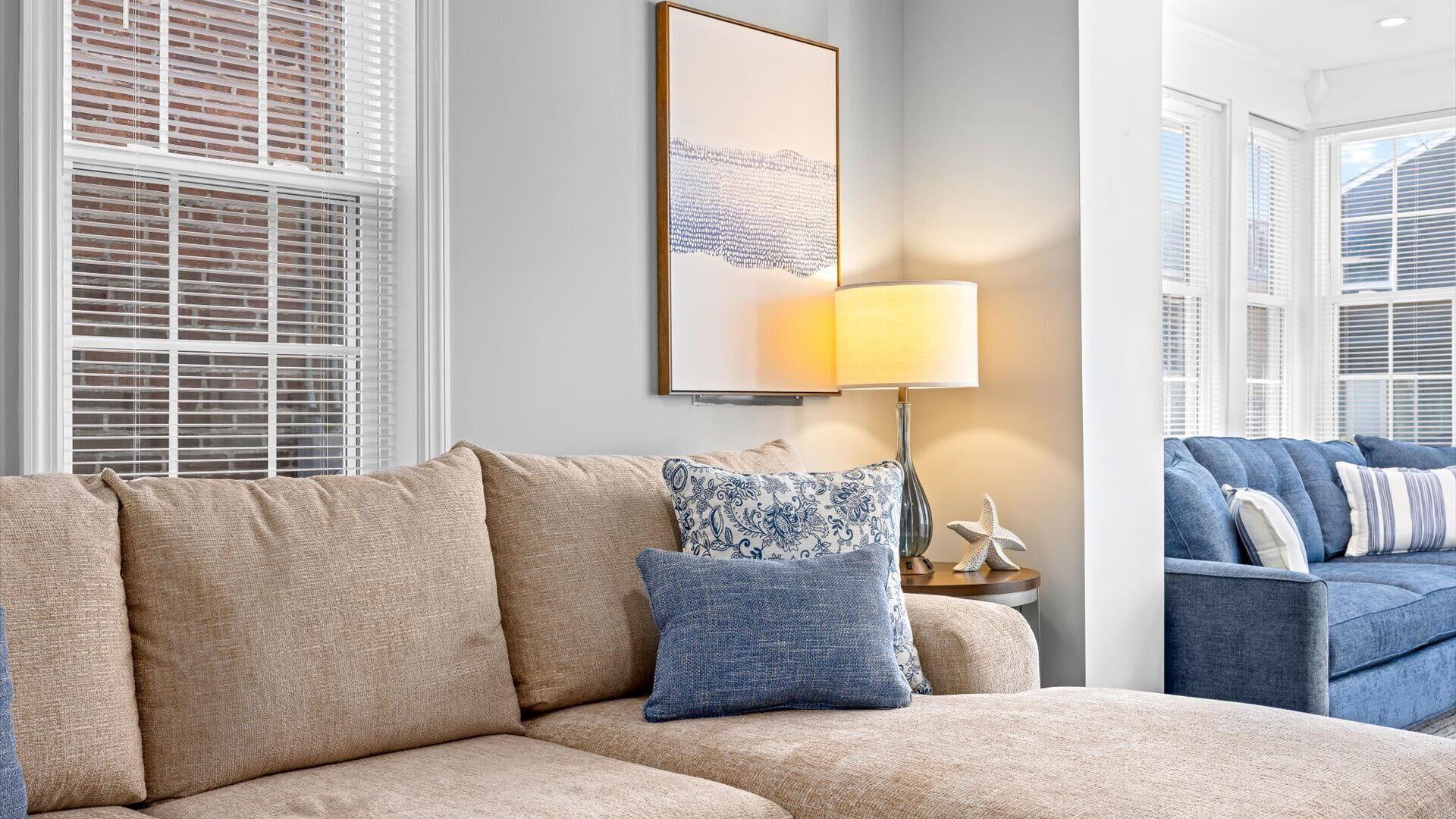 A living room with a beige sectional sofa, a patterned throw pillow, a wall-mounted lamp, and a framed artwork.