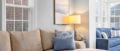 A living room with a beige sectional sofa, a patterned throw pillow, a wall-mounted lamp, and a framed artwork.