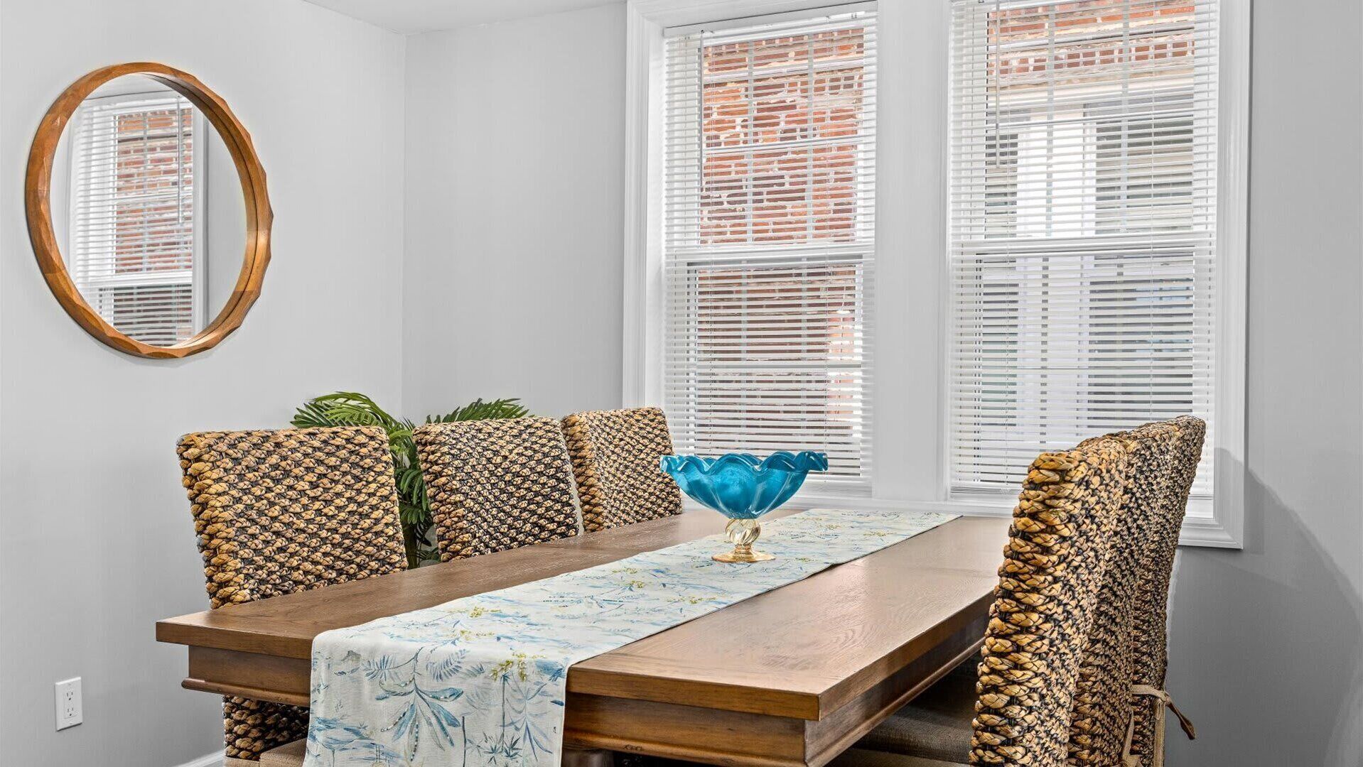 A dining area with a wooden table, patterned chairs, a blue glass bowl centerpiece, and white blinds on windows.