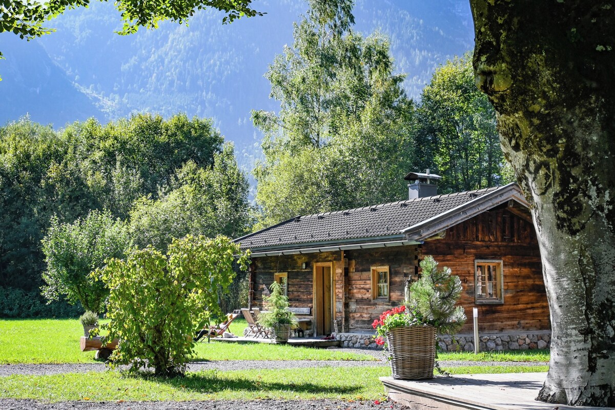 Une cabane rustique en rondins de bois, avec des fondations en pierre, une cheminée et une véranda dotée de sièges extérieurs.