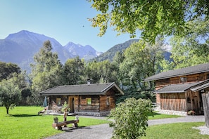 Une cabane en bois rustique, posée sur des fondations en pierre, entourée d’une végétation luxuriante et avec des montagnes en toile de fond.