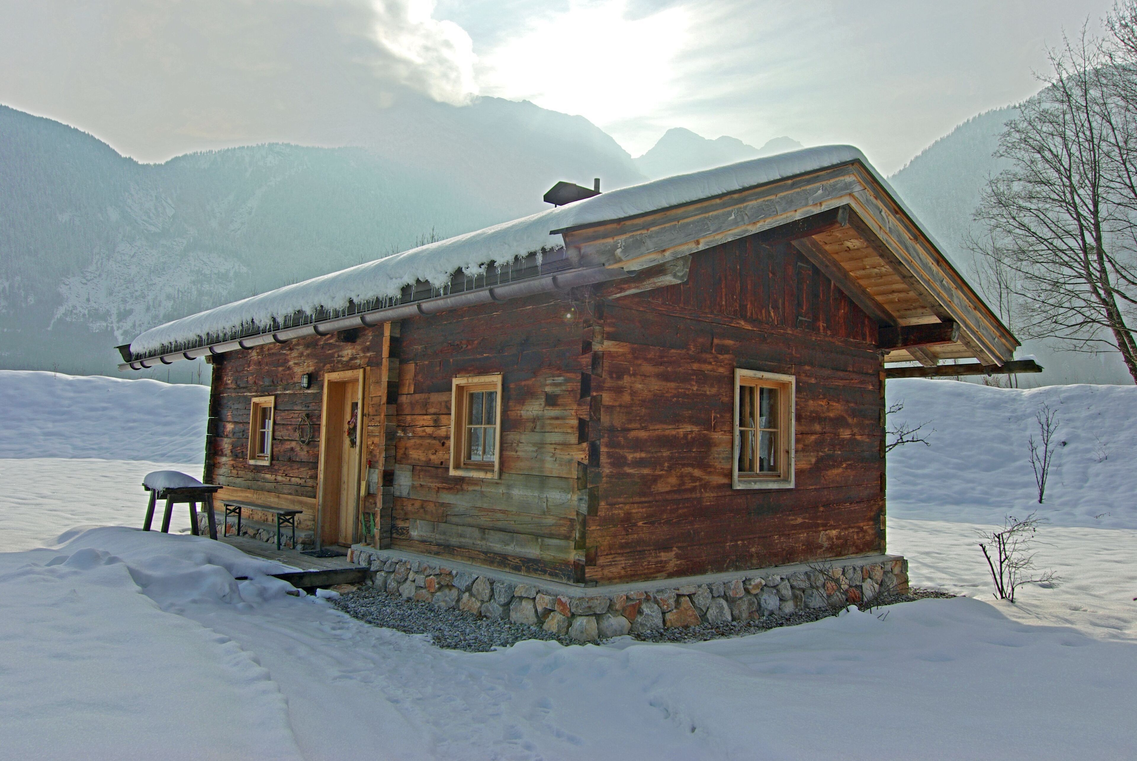 A wooden cabin with a stone foundation, wooden walls, and a snow-covered roof, surrounded by snow and mountains.