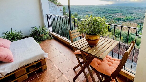 A balcony with a wooden table, chairs, a potted plant, and a bed with pillows.