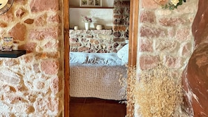 A stone-walled room with a bed, a shelf with books, and a potted plant.