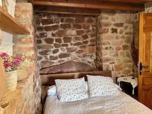 A stone-walled bedroom with a wooden headboard, a bed with patterned bedding, and a small table with a woven tablecloth.