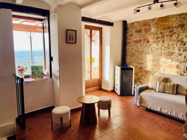 A cozy room with a stone wall, a sofa, a small table, a stool, a refrigerator, and a balcony with a view.