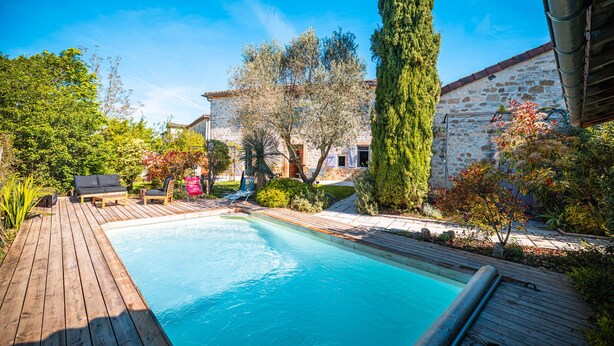 Um deck de madeira com piscina, rodeado por vegetação e um edifício de pedra.