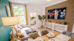 A modern living room with a dining area, a TV mounted on a wooden panel wall, and a view into the kitchen. - Urban Haven Central Retreat | Free Parking | 16PPL (Sheffield)
