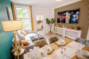 Ein modernes Wohnzimmer mit Essbereich, einem an einer Holzpaneelwand montierten Fernseher und Blick zur Küche.