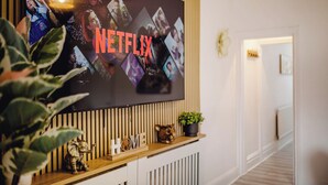 A wall-mounted television displaying the Netflix logo, a wooden shelf with decorative items, and a potted plant. - Urban Haven Central Retreat | Free Parking | 16PPL (Sheffield)