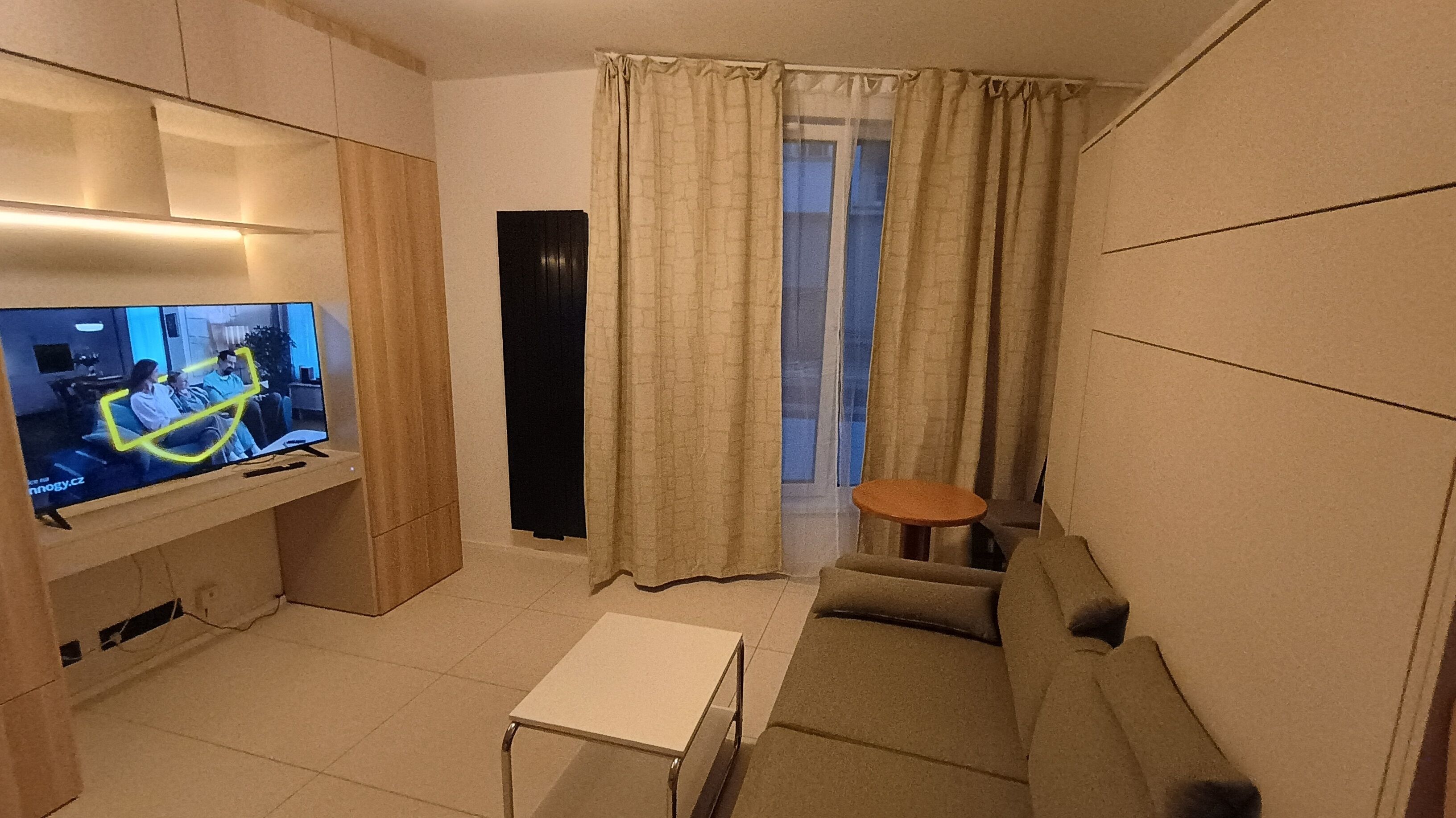 Een moderne woonkamer met een flatscreen televisie, een bank, een salontafel en een raam met gordijnen.