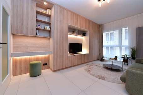 A modern living room with built-in shelves, a flat-screen TV, a round coffee table, and a green ottoman.