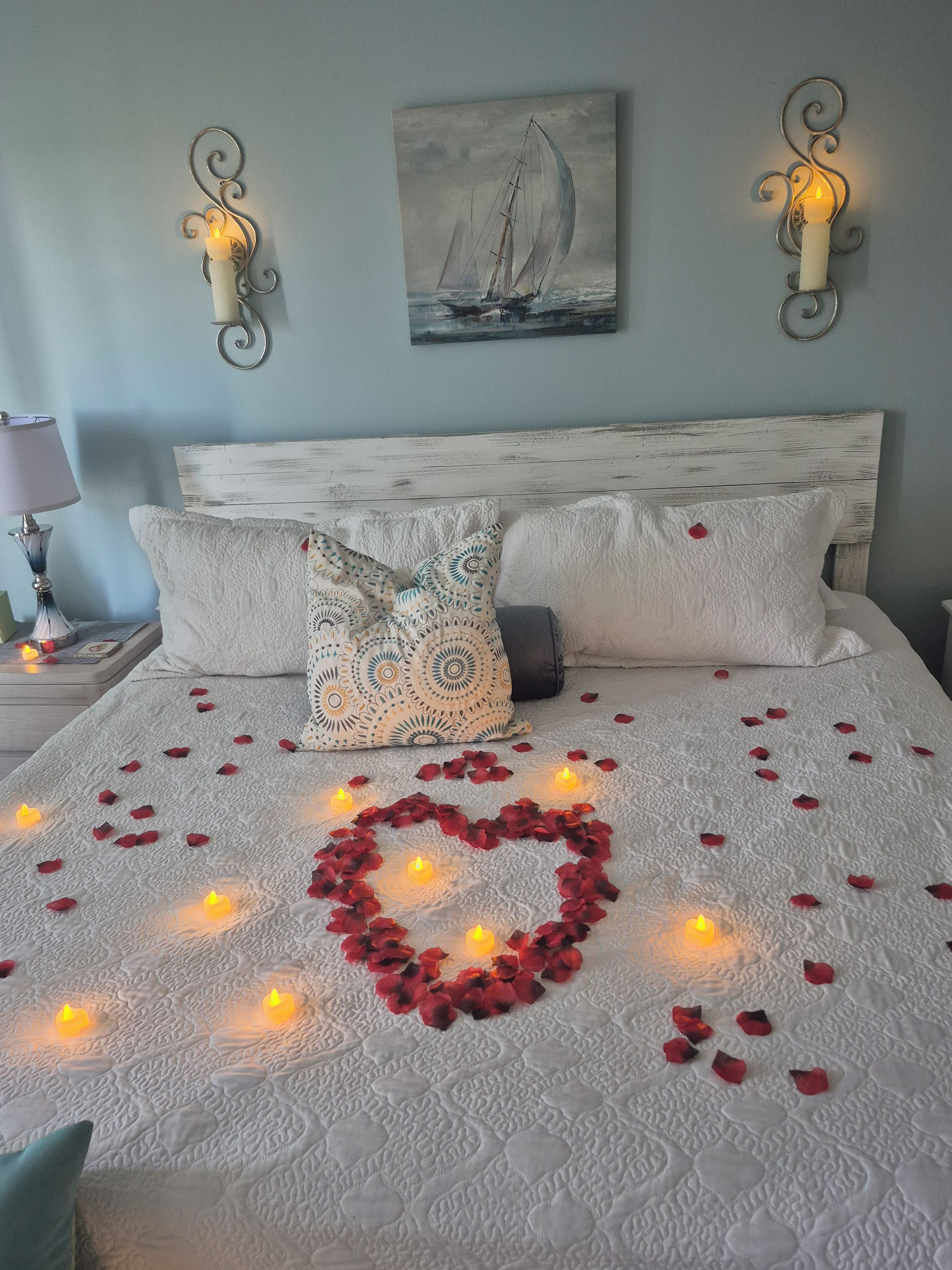A bed with a heart-shaped arrangement of rose petals and candles, two wall sconces, and a sailboat painting on the wall.