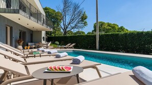 A modern pool area with lounge chairs, a table with a watermelon slice, and a view of a building with a balcony.