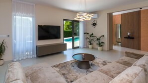 A modern living room with a flat-screen TV, a coffee table, and a sofa. There is a sliding glass door leading to a pool area.