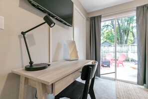 Ein Zimmer mit Schreibtisch, Stuhl, Lampe, Laptop und einer Glastür, die zu einem Außenbereich mit Sitzgelegenheiten führt.