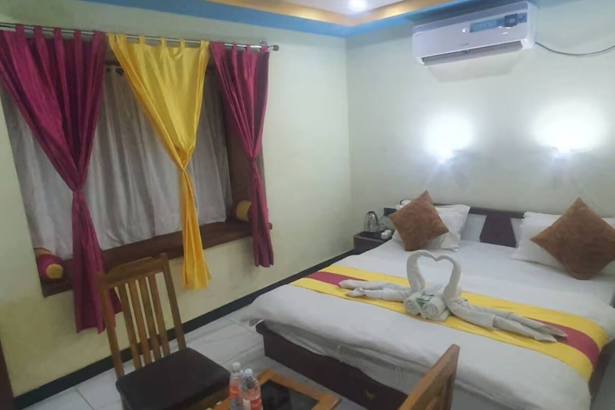 A hotel room with a bed, a chair, a small table with bottles, curtains, and an air conditioning unit.