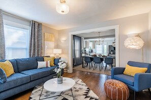 A modern living room with a sofa, coffee table, and ottoman, a dining area with chairs, and a kitchen in the background. - 21 - 2Bd Retreat Near KU Med 21 (Kansas City)