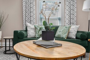 Ein grünes Sofa mit gemusterten Kissen, ein runder Couchtisch mit einem Buch und einer Topfpflanze sowie ein Fenster mit gemusterten Vorhängen.