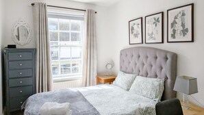 A bedroom with a bed, a dresser, a window with curtains, a ceiling fan, and framed botanical prints on the wall. - Chichester City Centre Flat -  a flat that sleeps 2 guests  in 1 bedroom (Chichester)