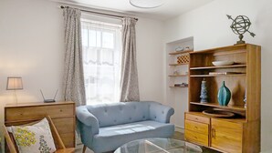 A living room with a blue armchair, a glass coffee table, a wooden cabinet with decorative items, and a window with patterned curtains. - Chichester City Centre Flat -  a flat that sleeps 2 guests  in 1 bedroom (Chichester)
