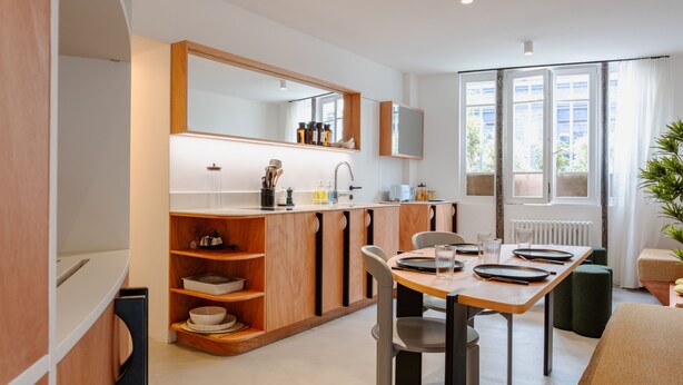 A modern kitchen with wooden cabinets, a dining table with chairs, and a large window providing natural light. - Babylon Montmartre (Paris)